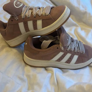 Adidas Campus sneakers i beige mocka - Säljer ett par Adidas Campus sneakers i beige mocka med klassiska vita ränder på sidorna och Adidas logga på plös och häl. Skorna har rund tå, platt sula och breda snören med zigzag-mönster i beige och vitt. Perfekt för dig som gillar retrovibe och snygga detaljer.