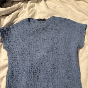Blå strukturerad topp från Zara - Säljer en blå topp från Zara i storlek S. Toppen har en cool strukturerad yta och rund hals. Modellen är kortärmad och har en slim passform som sitter snyggt på kroppen. Perfekt att styla med jeans eller kjol för en fräsch look.