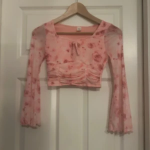 Rosa blommig croppad mesh-top - Supersöt croppad mesh-top i ljusrosa med blommigt mönster och långa trumpetärmar. Toppen har en snygg knytning framtill och lätt rynkade detaljer över magen. Perfekt för dig som gillar romantisk och trendig stil.