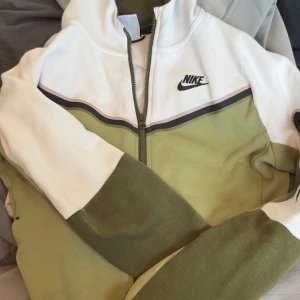 Nike zipjacka i vitt och olivgrönt - Nike zipjacka i vitt och olivgrönt med coolt svart logotryck på bröstet. Jackan har hel dragkedja framtill, kontrastpaneler och en praktisk ficka med dragkedja på ärmen. Perfekt för dig som gillar sportig och avslappnad stil.