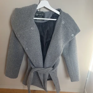 Grå kappa med huva  - Snygg grå kappa från Amisu med stor huva och knytskärp i midjan. Kappan har dubbelknäppning med knappar framtill och är helfodrad med svart innerfoder. Perfekt för dig som gillar stilren och modern look.