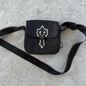 Trapstar side bag svart äkta  - Snygg svart axelväska från Thrasher med justerbar rem och coolt metallspänne i unik form på framsidan. Väskan har ett diskret rutmönster på materialet och en praktisk innerficka med dragkedja. Perfekt för att bära dina viktigaste grejer.