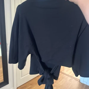 Svart omlottopp med knyt - Svart omlottopp med knyt framtill och trekvartsärm. Toppens stil är enkel och clean, med en croppad passform som ger en snygg siluett. Perfekt att matcha med höga jeans eller kjol för en chill och trendig look.