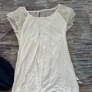 Vit topp med spetsärmar - Säljer en söt vit kortklänning med romantiska spetsärmar och rund halsringning. Klänningen har en mjuk och lätt känsla, perfekt för varma dagar. Spetsdetaljerna på axlarna ger en extra touch av stil och gör den superfin att bära.
