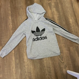 Grå Adidas hoodie med svarta ränder - Säljer en grå hoodie från Adidas i storlek L med den klassiska svarta treklövern och loggan på bröstet. Tröjan har svarta ränder längs ärmarna, huva och långa ärmar med muddar. Perfekt för chill eller träning, riktigt snygg streetstyle.