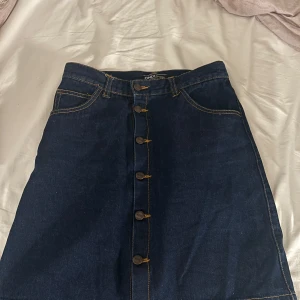 Mörkblå jeanskjol från ONLY W28 - Snygg mörkblå jeanskjol från ONLY med klassisk A-linjeform och kontrastsömmar. Kjolen har framfickor, bakfickor och knappar längs hela framsidan. Måtten ser man på sissta bilden