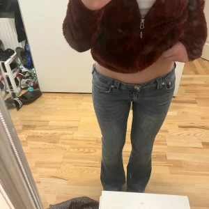 True Religion lågmidjade bootcut jeans - Säljer ett par blå jeans från True Religion med klassisk bootcut och låg midja. Jeansen har snygga slitningar, silverfärgade knappar och detaljerade bakfickor med rosa söm logga. Materialet är jeans med en skön passform som sitter loose över benen.