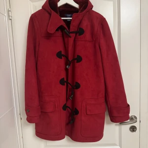 Röd duffeljacka med huva från Chaps - Chaps Vinterjacka i duffelstil med knäppning • Mjuk fuskpälsfoder • Fastsydd huva • Svarta knäppfästen i togglestil Färg: Highland Claret Röd Damstorlek M (Medium) Butikspris: ca 2 100 kr  Denna mjuka vinterjacka är tung och mysig för kalla klimat. Den är av fantastisk kvalitet och helt ny, oanvänd.  Ungefärliga mått: Byst: 56 cm tvärs över från armhåla till armhåla Längd: 79 cm från axel till nederkant.