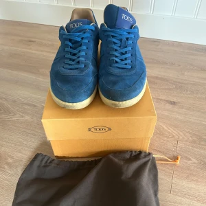 Blå mocka sneakers från Tod's - Snygga blå sneakers från Tod's i mjuk mocka med beige sula och klassisk snörning. Skorna har rund tå och coola gummidetaljer på hälen. Perfekta för dig som gillar stilrena och exklusiva sneakers med en unik touch. Nypris 450€