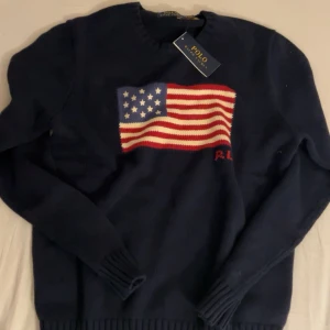 Mörkblå stickad tröja Polo Ralph Lauren - Snygg mörkblå stickad tröja från Polo Ralph Lauren med amerikansk flagga på bröstet. Tröjan har rund hals, långa ärmar och ribbade muddar. Perfekt för dig som gillar klassisk stil med en twist. Flaggan är stickad i rött, vitt och blått med RL-broderi. Bvsa! 