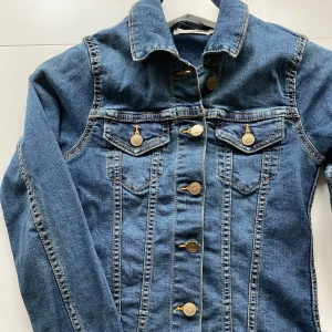 Blå jeansjacka från Black Shiny XS - Snygg klassisk blå jeansjacka från Black Shiny i storlek XS. Jackan har guldiga knappar framtill och på bröstfickorna, samt markerade sömmar som ger en cool look. Modellen är normal i passningen och har långa ärmar. Perfekt att slänga över en t-shirt för en chill stil.