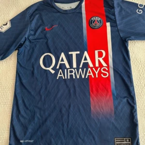 PSG Mbappé matchtröja Nike blå - Snygg PSG fotbollströja med Mbappé och nummer 7 på ryggen. Tröjan är marinblå med en röd vertikal rand och vita detaljer, tillverkad i lätt och ventilerande material. Qatar Airways-logga på bröstet och PSG-emblem. Perfekt för dig som älskar fotboll och Paris Saint-Germain.