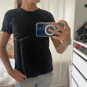 Svart t-shirt med silvriga nitar Zara - Svart t-shirt från Zara med rund hals och korta ärmar. På bröstet finns en ficka i siden tyg som är dekorerad med silvriga fyrkantiga nitar längs kanten. Materialet är mjuk bomull och fickan har en snygg kontrast mot resten av t-shirten.