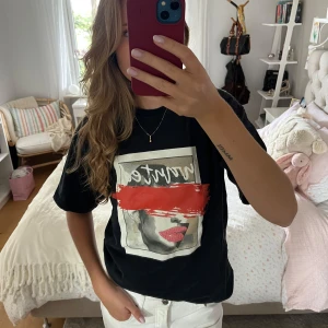 Svart t-shirt med grafiskt tryck - Svart t-shirt med coolt grafiskt tryck på framsidan som visar ett ansikte med röda läppar och en bred röd penseldrag över ögonen. Trycket har en vit ram och text ovanför. 