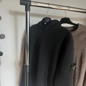 Svart hoodie från Stone Island - Svart hoodie från Stone Island med klassisk huva och stor magficka. Tröjan har den ikoniska Stone Island-loggan på vänster ärm och är gjord i mjuk bomull. Perfekt för dig som gillar streetwear och vill ha en clean look.