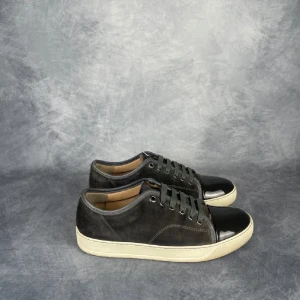 Svarta sneakers från Lanvin med lackad tå - Snygga svarta sneakers från Lanvin med cool lackad tå och ovandel i mjuk mocka. Skorna har gråa snören, rund tå och vit platt sula som ger en clean vibe. Perfekt för dig som gillar stilrena detaljer och exklusiv känsla.