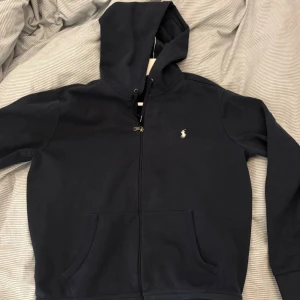 Marinblå hoodie från Ralph Lauren - Snygg marinblå hoodie från Ralph Lauren med dragkedja och klassisk Polo-logga broderad på bröstet. Tröjan har huva med snörning, två fickor fram och långa ärmar. Perfekt för chill dagar eller när du vill ha en clean look. 