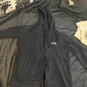 Svart hoodie från EA7 Emporio Armani - Svart hoodie från EA7 Emporio Armani med dragkedja och huva med snörning. Stilren logga på bröstet och långa ärmar. Perfekt för chill dagar eller när du vill ha en enkel men snygg look. Mjuk bomullskänsla och klassisk passform.