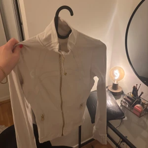 Vit zipjacka från Lululemon - Snygg vit zipjacka från Lululemon med guldfärgade dragkedjor och figurnära passform. Jackan har hög krage, långa ärmar och två fickor med dragkedja. Perfekt för träning eller chill, tillverkad i mjuk polyesterblandning.