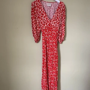 Röd blommig långklänning Feather and Bone - Säljer en superfin långklänning från Feather and Bone London med röd och ljusrosa blommigt mönster. Klänningen har v-ringning, puffiga trekvartsärmar och knyt i ryggen för extra snygg passform. Materialet är mjukt och lätt, perfekt för vår och sommar.