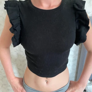 Svart croppad topp med volangärmar från Zara - Supersnygg svart croppad topp från Zara med ribbad struktur och stora volangärmar som ger en cool och trendig vibe. Perfekt att styla med höga jeans eller kjol för en riktigt chic look. Materialet är mjukt och stretchigt, vilket gör den bekväm att bära hela dagen.