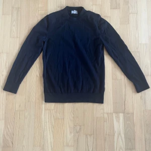 Svart polotröja i bomull - Snygg svart polotröja med lång ärm och ribbade muddar. Tröjan har en enkel och stilren design med rundad hals och är gjord i mjuk bomull som känns skön mot huden. Perfekt för dig som gillar minimalistisk stil och vill ha en tröja som funkar till många outfits.