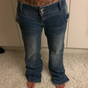 Blå lågmidjade baggy jeans från Junk de Luxe - Säljer ett par blå jeans  med låg midja och baggy passform. Jeansen har två knappar i midjan, slitningar och tvättade detaljer framtill och baktill, samt stora bakfickor med dekorativa sömmar. Perfekt för dig som gillar avslappnad stil. 