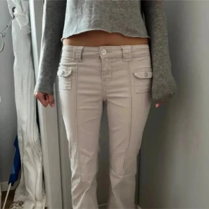 Beige bootcut byxor med fickor - Snygga beige bootcut-byxor med låg midja och coola fickdetaljer framtill. Byxorna har breda bälteshällor och knäppning med knapp och dragkedja. Passar dig som gillar en avslappnad men trendig stil. Perfekta till sneakers eller boots. 38/M