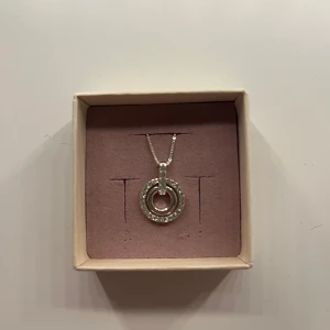 Silverfärgat halsband med cirkelhänge - Stilrent halsband i silverfärg med ett cirkelformat hänge dekorerat med gnistrande stenar runt om. Kedjan är tunn och smidig, perfekt för att ge en elegant touch till din look. Hänget har en modern design med dubbla ringar och små stenar som fångar ljuset snyggt.