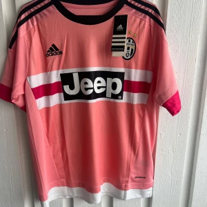 Juventus Dybala rosa matchtröja Adidas - Juventus fotbollströja med Dybala 21 på ryggen, tillverkad av Adidas i lätt och ventilerande climacool-material. Tröjan är rosa med svarta och vita detaljer, har svarta Adidas-ränder på axlarna och Jeep-logga på bröstet. Kortärmad med Serie A-märke på ärmen.