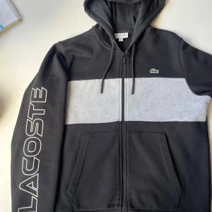 Svart och grå hoodie från Lacoste - Snygg svart hoodie från Lacoste med bred grå panel över bröstet och klassisk krokodillogga. Stor LACOSTE-text på ena ärmen, huva med snörning och dragkedja framtill. Mjuk bomullskänsla och praktiska fickor. Perfekt för chill eller streetwear.