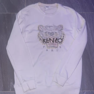 Vit sweatshirt med tiger från Kenzo - Kenzo Paris sweatshirt i vitt med ikonisk broderad tiger på bröstet och logotyp. Tröjan har rund hals, ribbade muddar och är långärmad. Perfekt för dig som gillar streetstyle och vill sticka ut med ett snyggt statement-plagg.