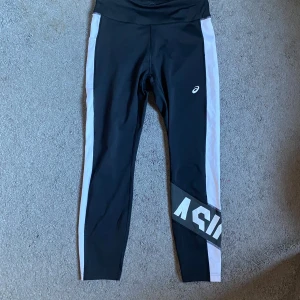 Svarta leggings från Asics med vita ränder - Snygga svarta leggings från Asics med vita ränder längs sidorna och en stor logga på ena benet. De har hög midja och är gjorda i ett stretchigt syntetmaterial som passar perfekt för träning. 