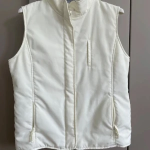 Vit väst S - Snygg vit väst från Xlind med hög krage och diskreta fickor med dragkedja. Västen har en stilren look och är perfekt att slänga över en hoodie eller tröja när det är kyligt ute. Passar dig som gillar enkel och clean stil.