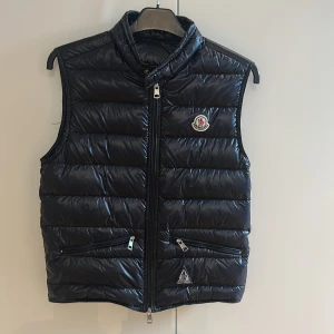 Svart dunväst från Moncler - Snygg svart dunväst från Moncler med glansig finish och klassisk logga på bröstet. Västen har ståkrage, två dragkedjefickor fram och quiltad design. Perfekt att slänga över en hoodie för en cool look. Dragkedja framtill och diskreta detaljer.