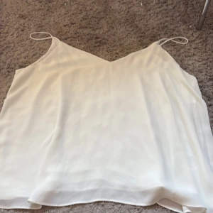 Vit linnetopp med tunna axelband - Säljer en stilren vit linnetopp med tunna axelband och v-ringad hals. Toppen har en lätt och flowy passform, perfekt för varma dagar. Materialet känns luftigt och ger en fräsch look. Enkel att matcha med jeans eller kjol för en clean vibe.