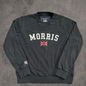 Svart sweatshirt från Morris med tryck - Svart sweatshirt från Morris i storlek medium med stort vitt Morris-tryck och en broderad brittisk flagga på bröstet. Tröjan har rund hals, ribbade muddar och är tillverkad i mjuk bomull. Perfekt för dig som gillar klassisk stil med en sportig touch.