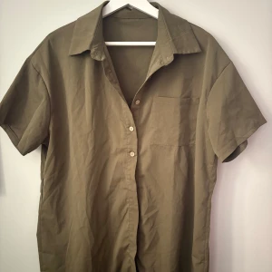 Olivgrön kortärmad skjorta med ficka - Snygg olivgrön skjorta med korta ärmar och klassisk krage. Skjortan har knappar framtill och en bröstficka som ger en chill vibe. Materialet känns mjukt och luftigt, perfekt för varmare dagar. Passar dig som gillar enkel och stilren look.