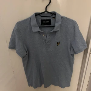 Ljusblå Lyle & Scott piké t-shirt - Fräsch ljusblå piké från Lyle & Scott med klassisk krage och två knappar framtill. T-shirten har korta ärmar och den ikoniska gula fågeln broderad på bröstet. Perfekt för en chill och stilren look. Materialet är mjuk bomull. Storlek S