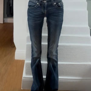 Mörkblå miss me bootcut jeans med låg midja  - Snygga blå jeans från Miss Me med bootcut passform och låg midja. Baksidan har två fickor med grymma paljettdetaljer och broderier som verkligen sticker ut. Jeansen har klassisk femficksdesign och slitningar framtill för en cool look. Passar som storlek 34/xs. 