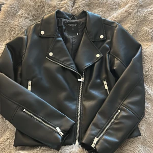 Svart skinnjacka från The Black Label - Snygg svart skinnjacka från The Black Label Collection i storlek M. Jackan har klassisk bikerstil med silvriga dragkedjor, bred krage med tryckknappar och flera fickor med zip. Perfekt för dig som vill ha en edgy look och gillar detaljer.