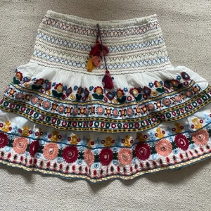Broderad vit kortkjol med volang - Superfin vit kortkjol med broderade blommor i rött, rosa, blått och gult. Kjolen har volanger, snörning med tofsar och färgglada detaljer som gör den unik. Perfekt för dig som gillar boho-stil och vill sticka ut med färg och mönster.