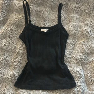 Svart ribbad linnetopp från Nelly - Svart ribbad linnetopp från Nelly i storlek S. Toppen har smala axelband och är figurnära med en enkel, stilren design. Materialet är mjukt och stretchigt, perfekt för att matcha med jeans eller kjol. Snygg att bära som den är eller under en skjorta.