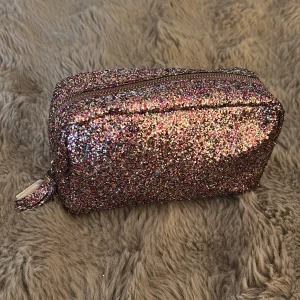 Glittrig neccesär i regnbågsfärger - Säljer en superglittrig neccesär med regnbågsfärgat glitter över hela ytan. Neccesären har en rektangulär form och stängs med dragkedja i metallic. Perfekt för dig som vill sticka ut och addera extra färg till din look!