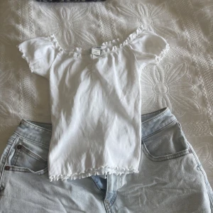 Vit ribbad topp från H&M - Säljer en söt vit ribbad topp från H&M med korta puffärmar och volangkant vid halsringning och nederkant. Toppen har en feminin och somrig stil, perfekt till jeans eller kjol.