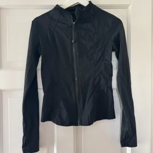 Svart tunn träningsjacka med dragkedja - Säljer en svart, tunn och smidig jacka med hel dragkedja framtill. Jackan har lång ärm och en kroppsnära passform. Jättesnygg som liknar lululemon