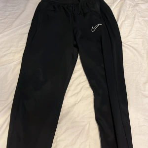 Svarta Dri-Fit träningsbyxor från Nike - Säljer ett par svarta träningsbyxor från Nike med Dri-Fit material. Byxorna har dragsko i midjan och en vit Nike-logga på ena benet. Perfekta för träning eller chill. Snygg och enkel design med normal passform.