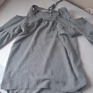 Randig offshoulder blus med knytband - Säljer en svartvit randig blus med offshoulder-design och knytband vid halsen. Blusen har långa ärmar och volangdetaljer upptill. Perfekt för dig som gillar en trendig och luftig stil.