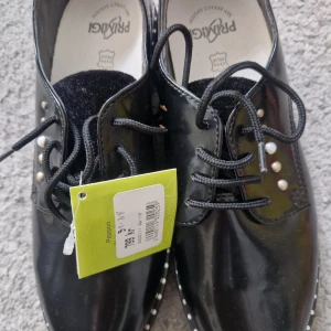 Svarta lackade skor med pärldetaljer från Primigi - Snygga svarta lackskor från Primigi med snörning och dekorativa pärlor på sidan. Skorna har rund tå, platt sula och markerade sömmar. Perfekta för dig som vill ha en stilren men ändå unik look.