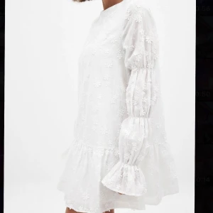 Vit blommig klänning med volangärm - Superfin vit klänning med blommigt broderi, volangdetaljer vid ärmslut och kjol, samt lång ärm. Öppen rygg med knytband upptill. Perfekt för sommarens tillfällen!